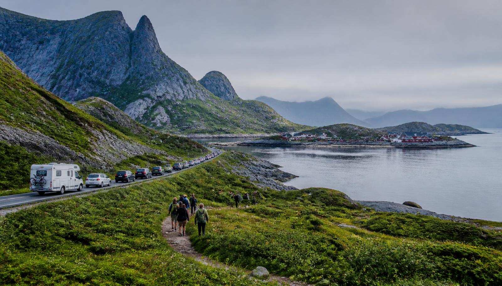 A queue of cars and a camper van along a narrow Lofoten coastal road, with hikers on a worn footpath and a fishing village across the fjord.